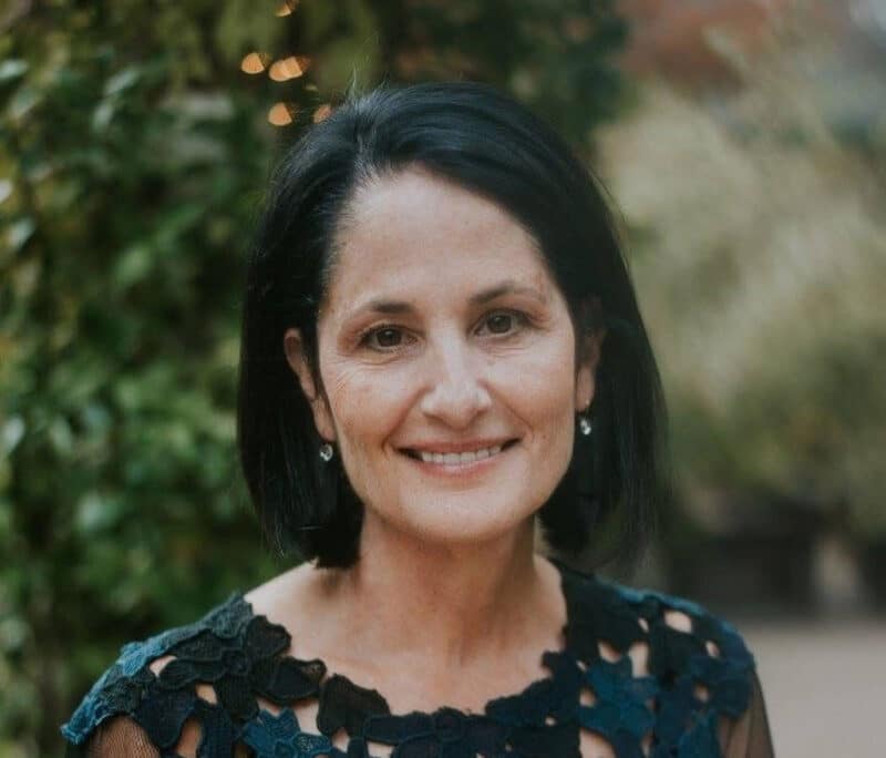 Alameda Post - Nancy Lee Bronstein, a woman with a black bob hairstyle, smiles at the camera in a beautiful headshot. She is in an elegant top that may be a dress, and in a garden.