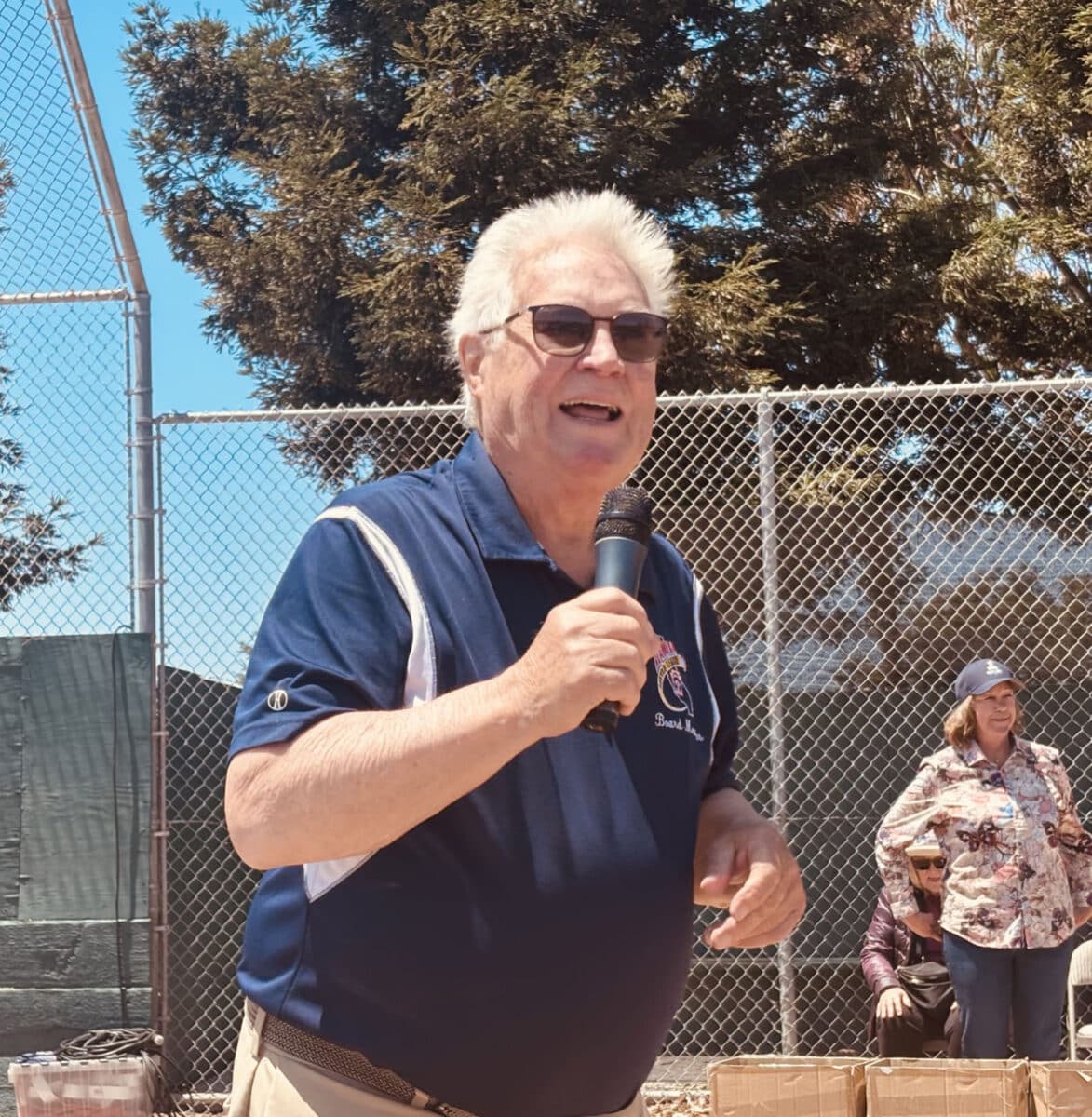 Alameda Post - Ron Matthews stands in front of a crowd at a baseball field holding a microphone and speaks.