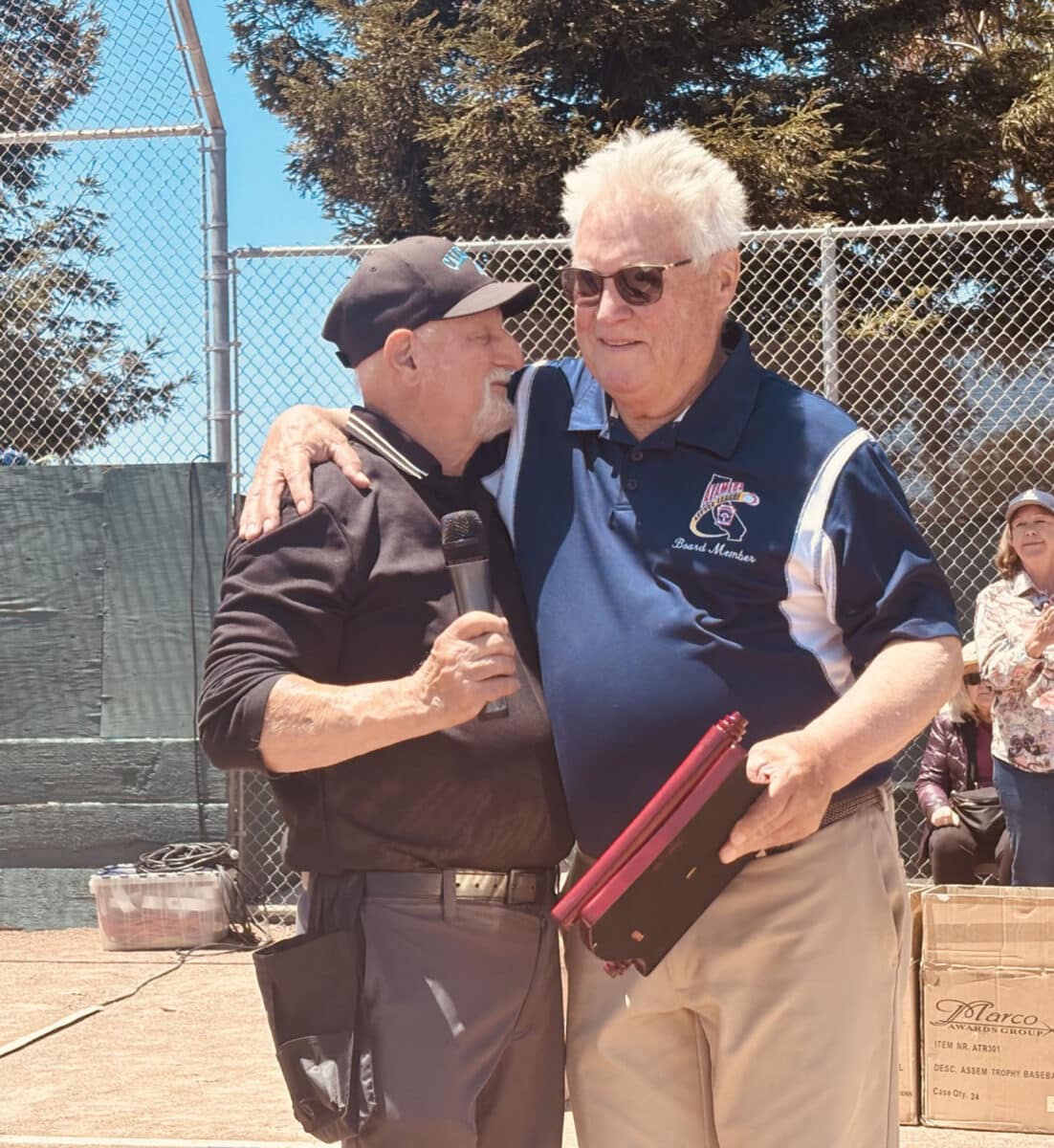 Alameda Post - Little League President Ron Matthews hugs another man in front of a crowd at the ball field. 