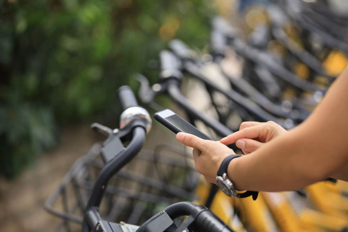 Alameda Post - A person uses their smartphone while standing next to a bike rack full of bike share bicycles.