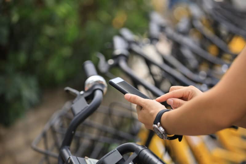 Alameda Post - A person uses their smartphone while standing next to a bike rack full of bike share bicycles.