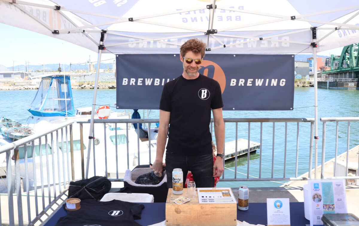 Alameda Post - A man stands behind the table at a booth. He is by the waterfront, and the sign says Brewbilt Brewing. 