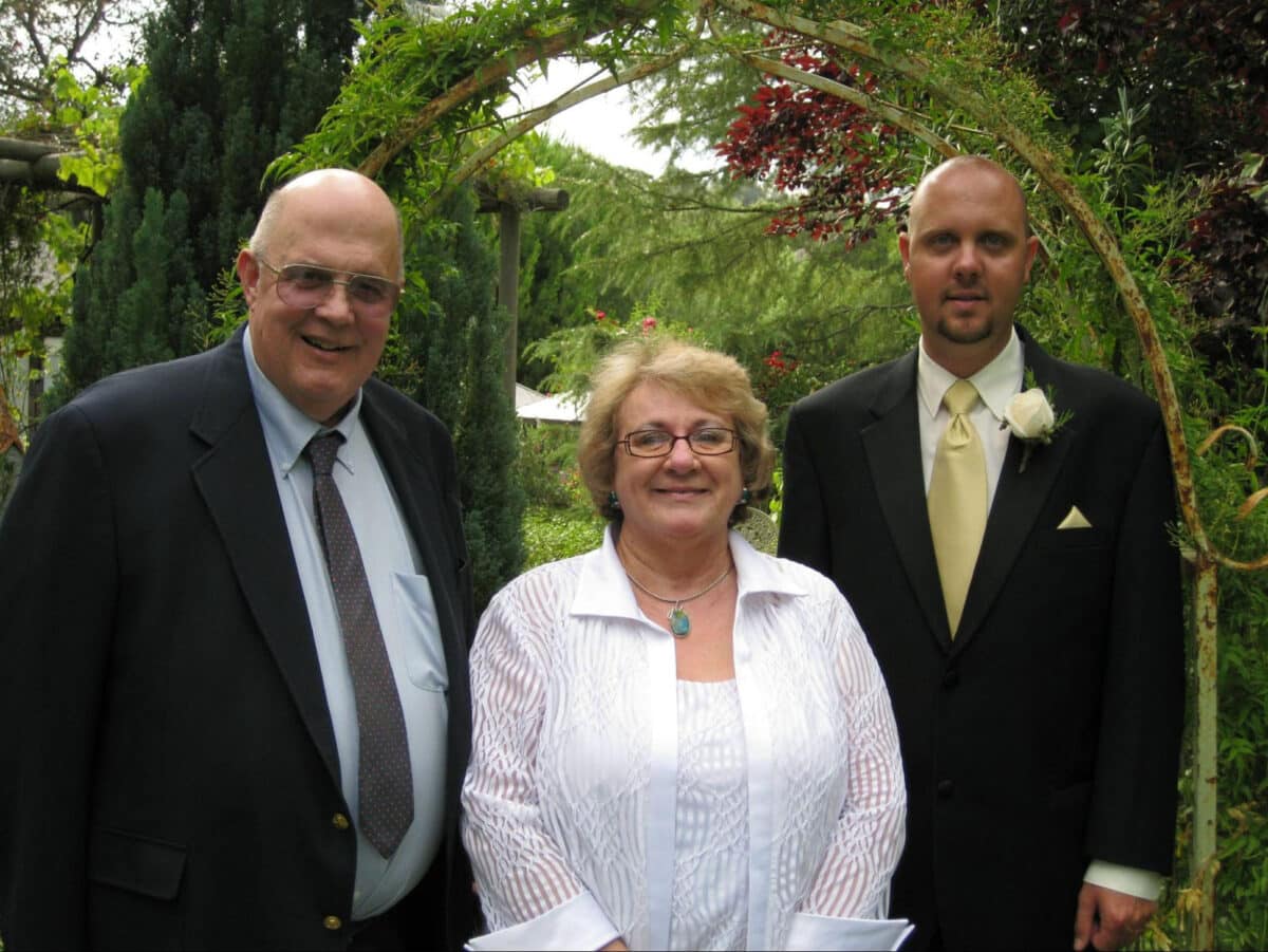 Alameda Post - Sandra Jo Amstutz stands in between two men. They are all dressed formally, and family resemblance and age suggest that the men are her husband and son at her son's wedding. 