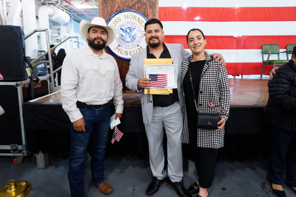 Alameda Post - Attendees of the USS Hornet's naturalization ceremony smile at the camera and hold American flags.