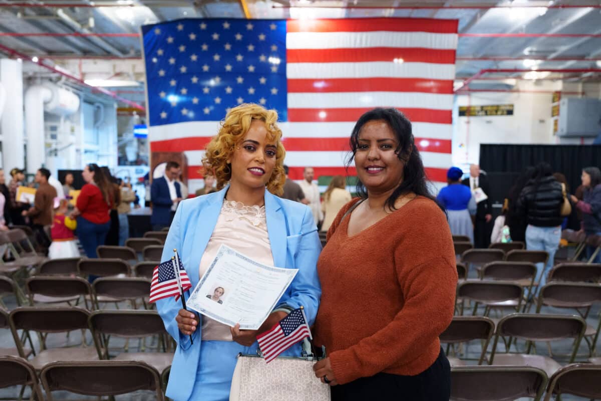 Alameda Post - Attendees of the USS Hornet's naturalization ceremony smile at the camera in front of the large American flag.