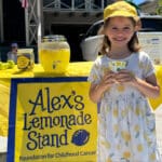 Alameda Post - A young girl wearing a dress with lemons on it stands in front of a lemonade stand. She holds a cup and smiles.