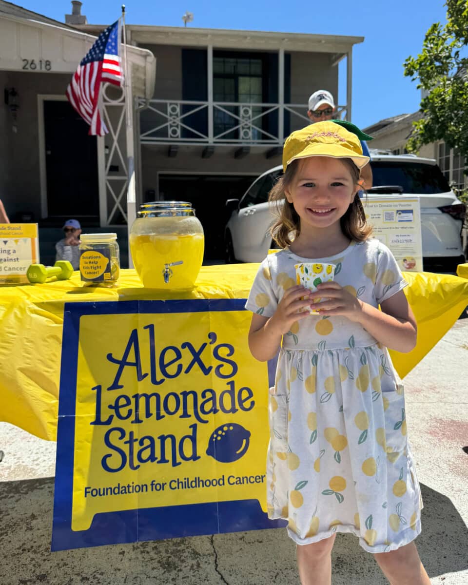 Alameda Post - A young girl wearing a dress with lemons on it stands in front of a lemonade stand. She holds a cup and smiles. 