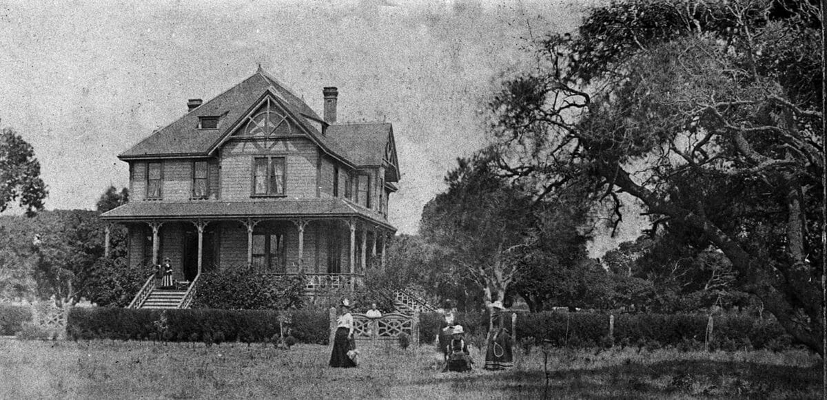 Alameda Post - A beautiful black and white photo of a family in front of a large victorian era home, hedges, hard, and trees. 