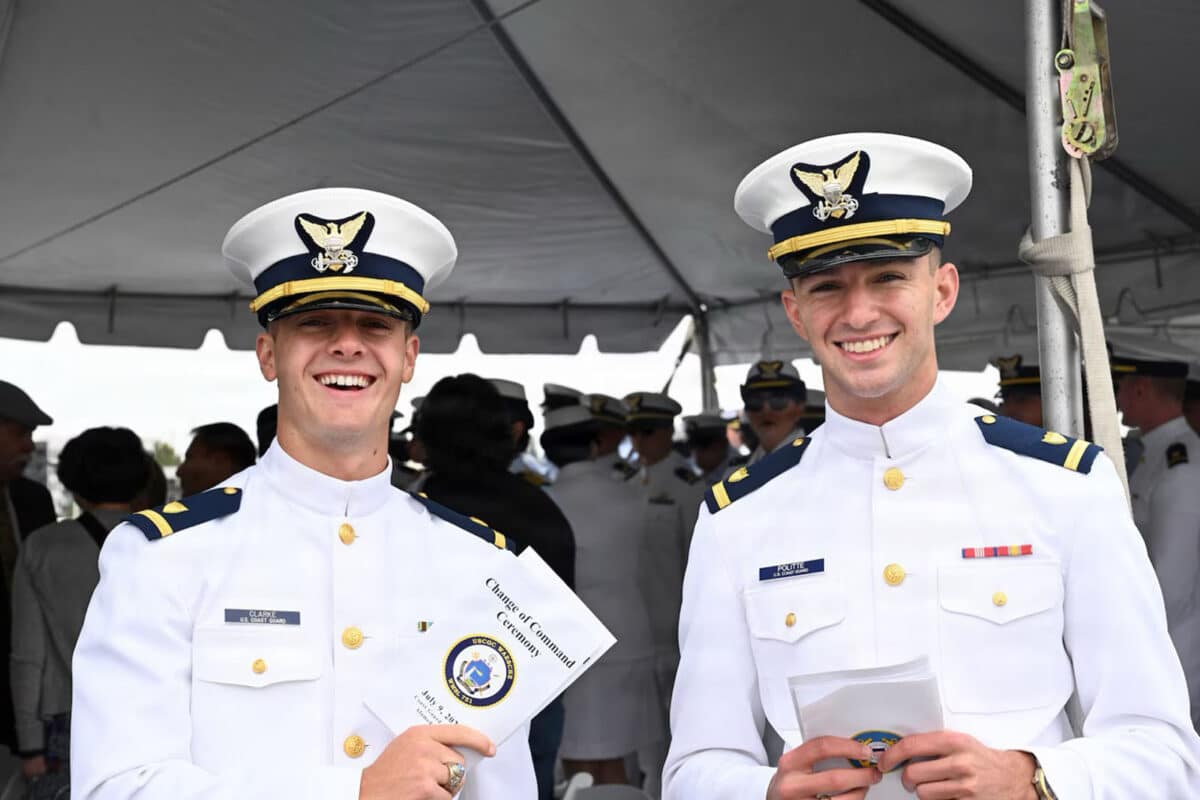 Alameda Post - Two young Coast Guard servicemen hold pamphlets for the Chance of Command Ceremony in front of a white event tent and smile. 