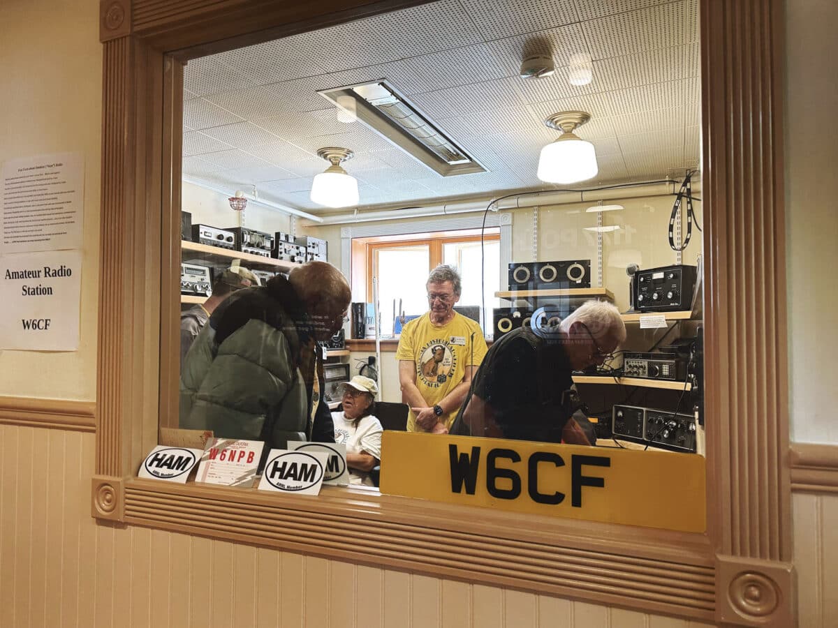 Alameda Post - A photo through a window into a recording booth. People are milling around and examining the area.