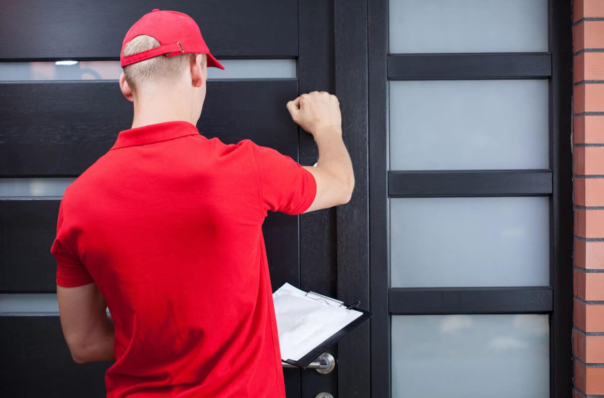 Alameda Post - A man in a red shirt and matching baseball cap holds a clipboard and knocks on a front door. 