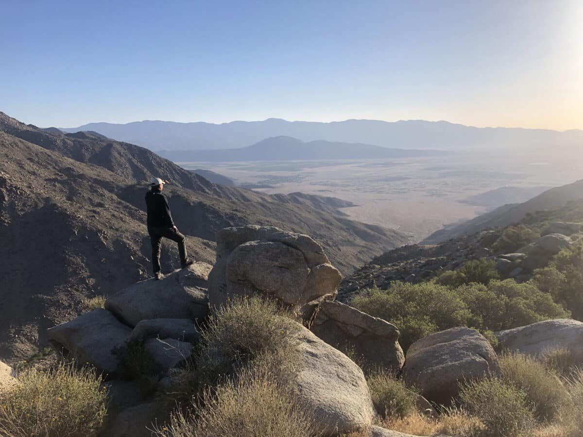 Alameda Post - A hiker stands with their food on a rock on the side of a large hill or mountain and looks out over the landscape. 