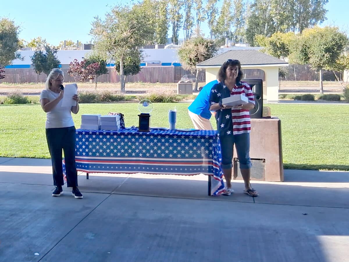Alameda Post - Two women stand at a table at the front of a pavilion. One speaks into a microphone and the other hold a box.