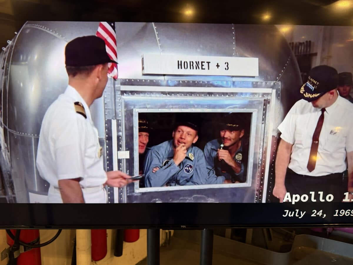 Alameda Post - Three astronauts smile and look through a window in a capsule. Two officers talk with them through the window.