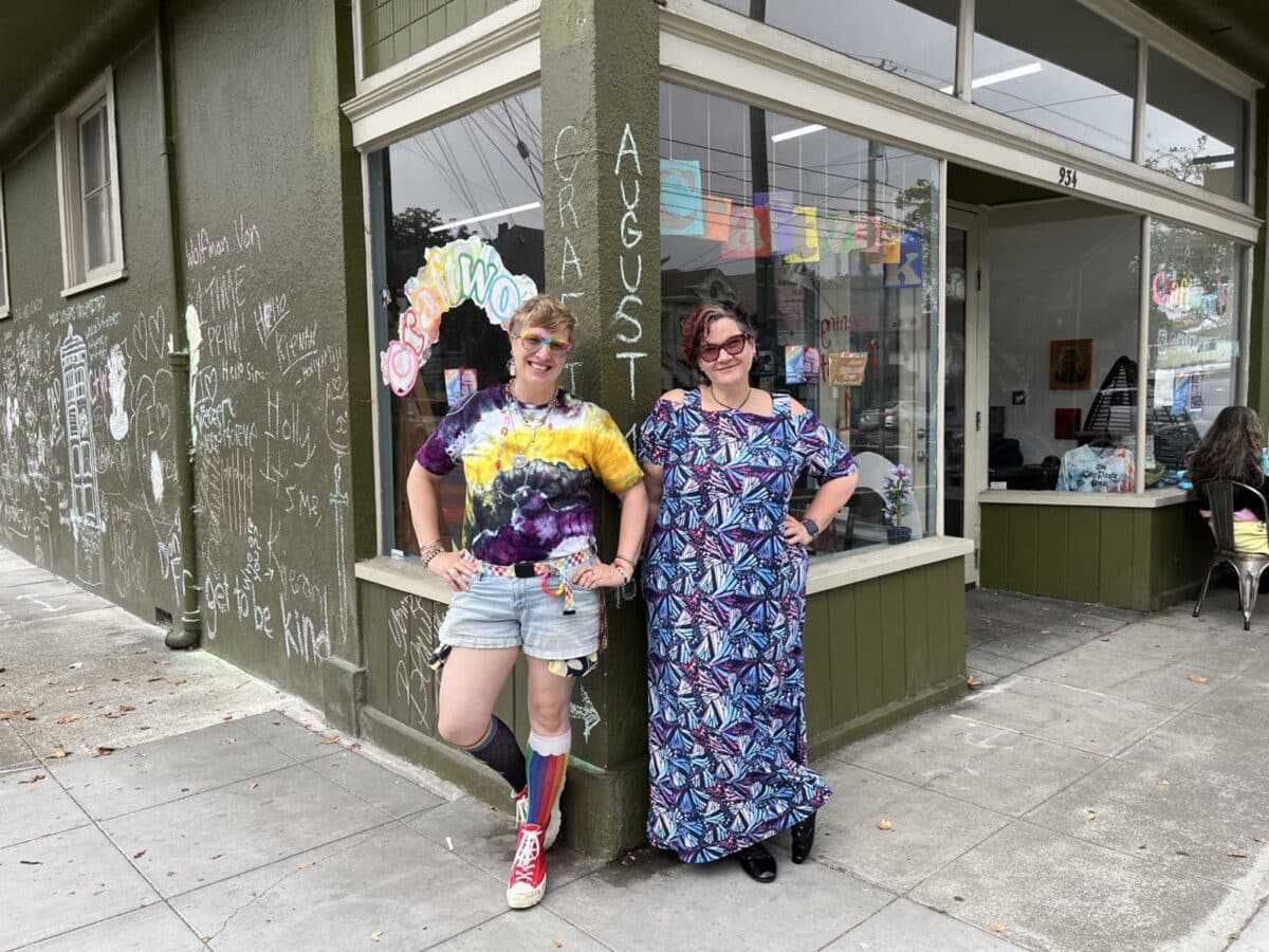 Alameda Post - Two of the owners of Craftwork stand at the corner of their store and smile. They have their hands on their hips and wear fun outfits.