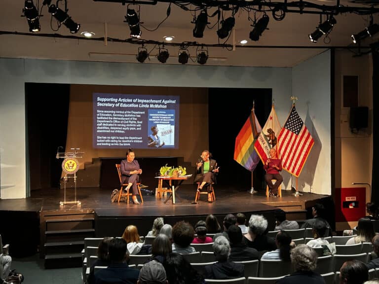 Alameda Post - Two women sit onstage and talk during a town hall. A slide deck is presented behind them, and the California, American, and Pride flags are on the stage.