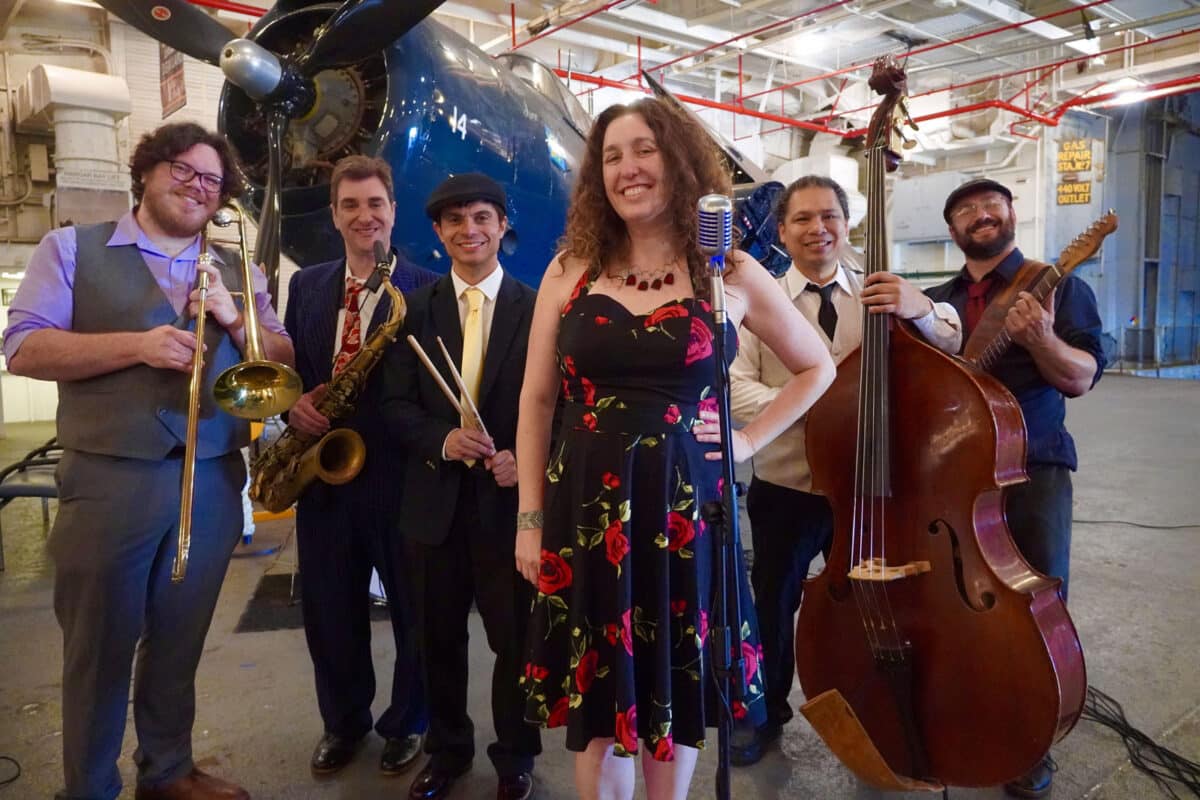 Alameda Post - The Death and Taxes Swing band pose and smile in front of a plane in a hanger or on a aircraft carrier. 