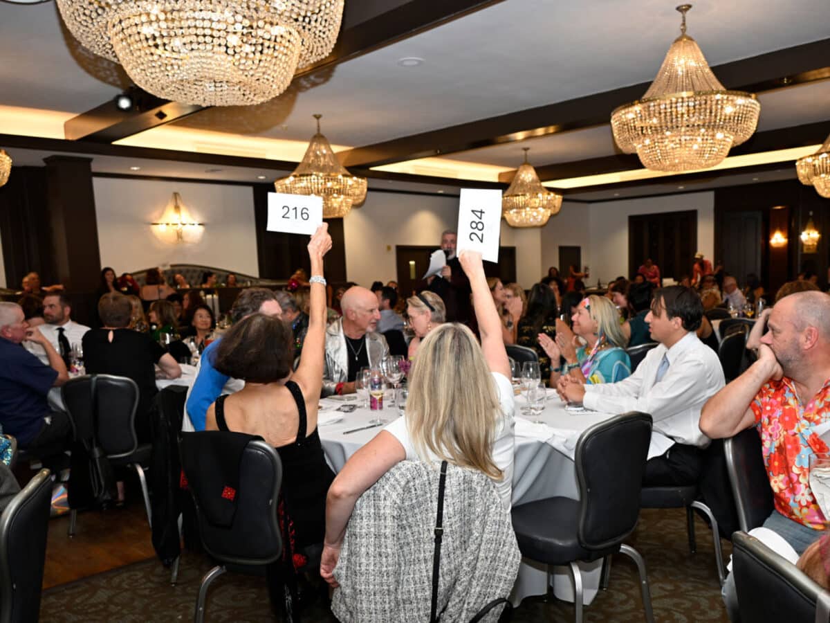 Alameda Post - A ballroom full of guests sitting at tables with white tablecloths. Some guests raise numbers into the air. 