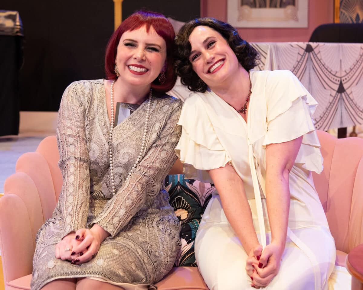 Alameda Post - Two women in Fallen Angels sit together on a sofa and smile while leaning towards each other and facing the audience. 
