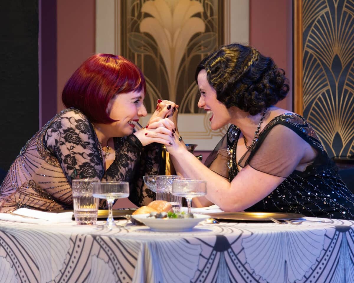 Alameda Post - Two actresses dressed in fabulous 1920s costumes and sitting at a well set table clasp hands, lean together, and giggle.