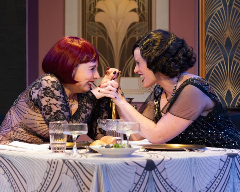 Alameda Post - Two actresses dressed in fabulous 1920s costumes and sitting at a well set table clasp hands, lean together, and giggle.