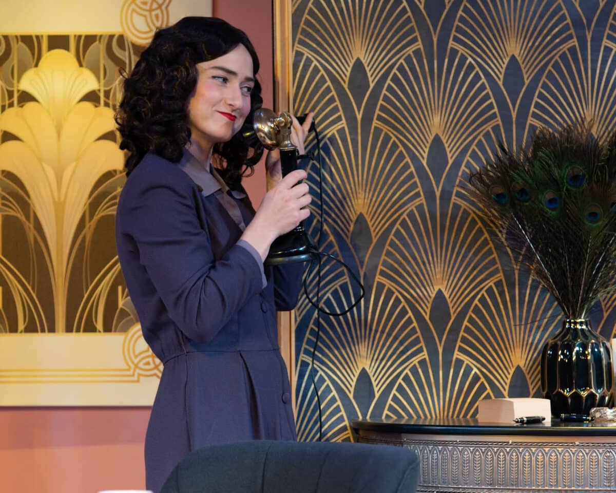 Alameda Post - In front of an art deco wall, a woman in a dark purple suit listens to an old fashioned telephone. 