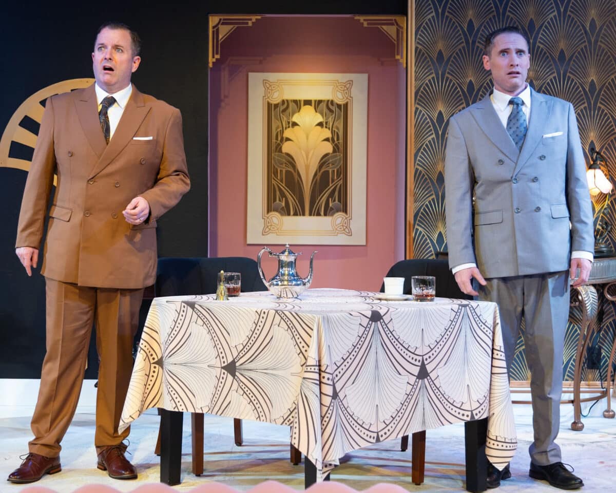 Alameda Post - Two men in suits stand onstage amongst a rich art deco set. 