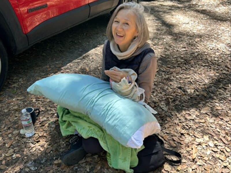 Alameda Post - A woman sits on the forest floor next to a car. She is gingerly holding her wrist and smiling at the camera.
