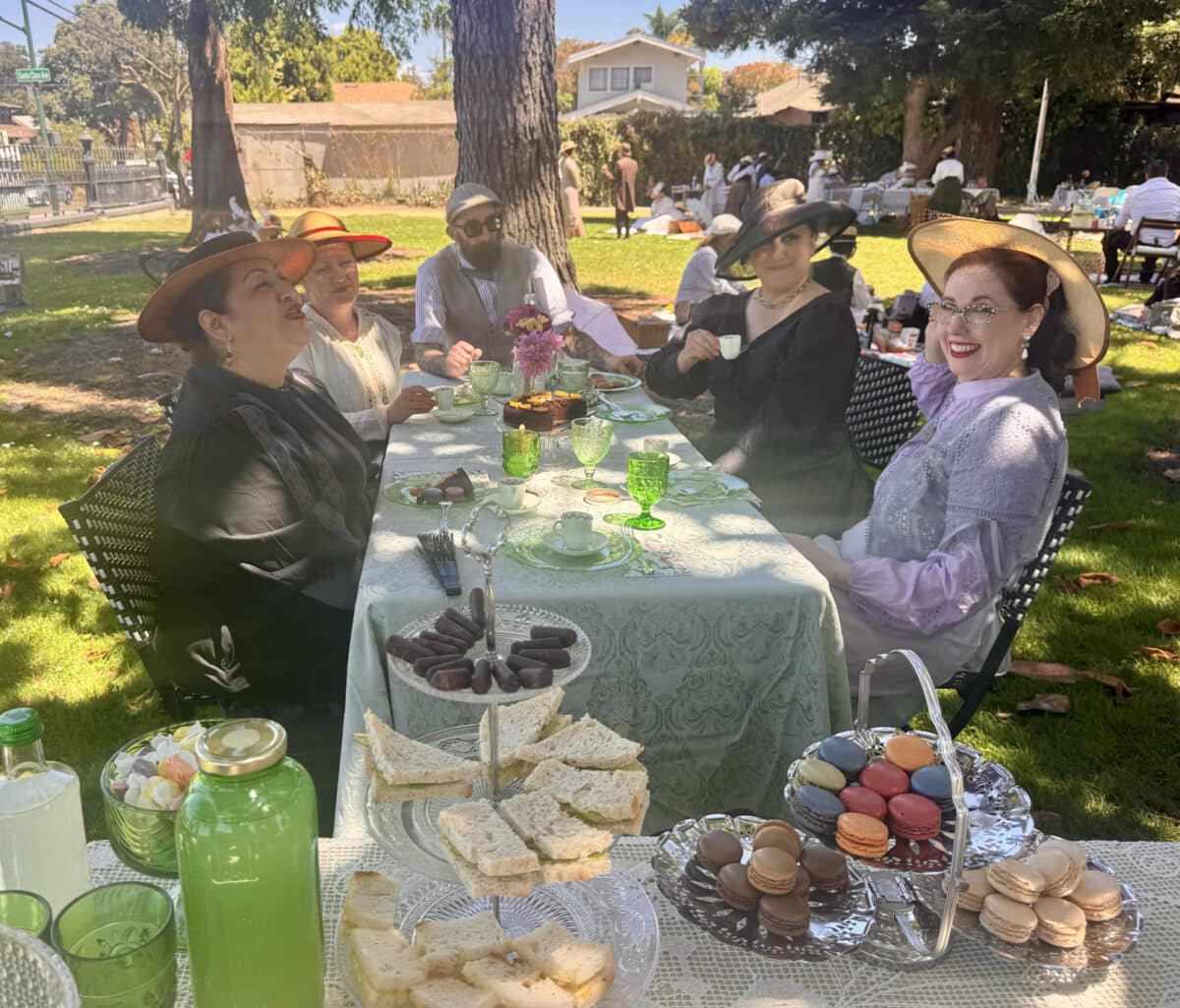 Alameda Post - A group of adults in Edwardian costumes sit outside at a table with a tablecloth and enjoy a picnic. 