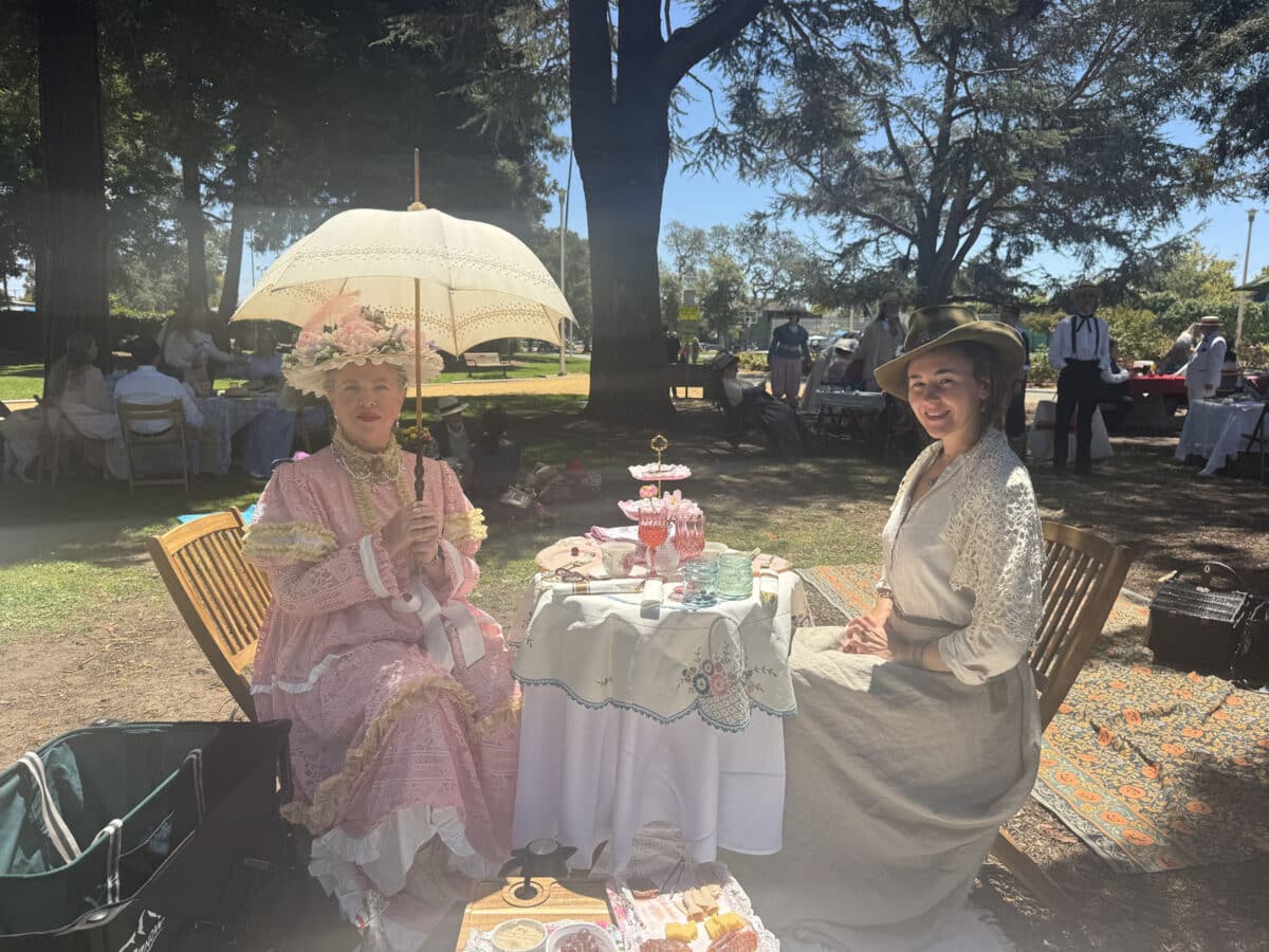 Alameda Post - Two women sit at a small, round, and well decorated table. One woman holds a parasol, and they are both in Edwardian garb. 