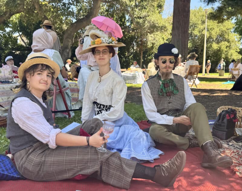 Alameda Post - Three young adults wearing Late Victorian or Edwardian costumes sit on a picnic blanket outdoors at Lincoln Park.