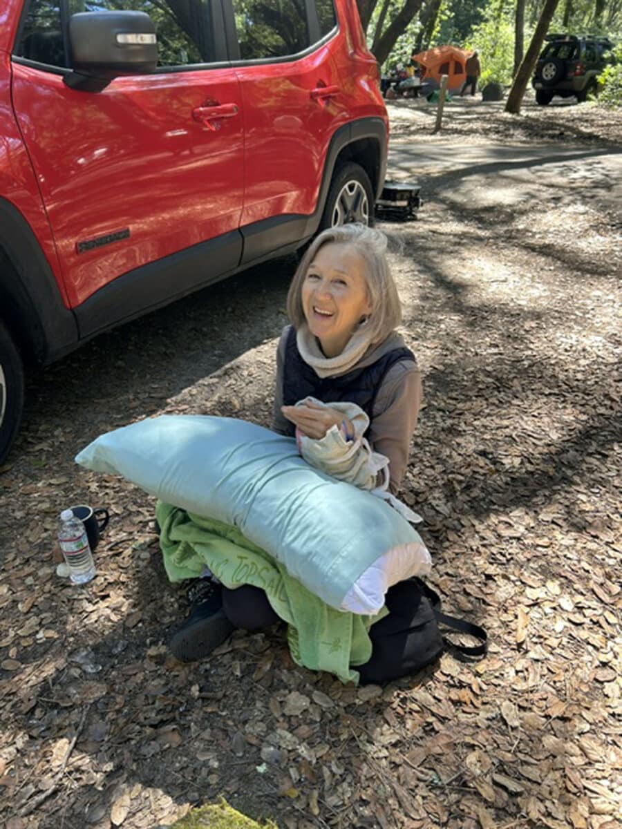 Alameda Post - A woman sits on the forest floor next to a car. She is gingerly holding her wrist and smiling at the camera.