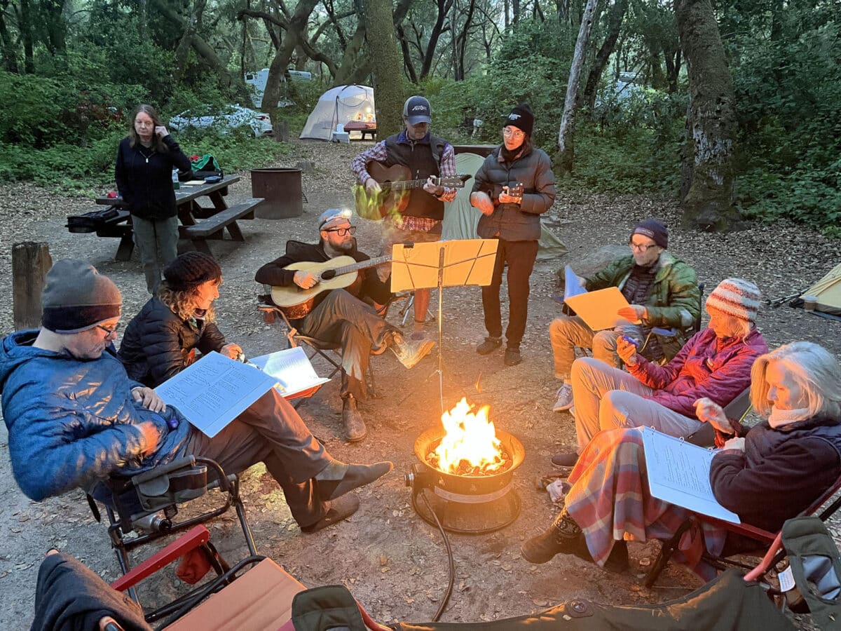 Alameda Post - A group of nine people sit and stand around a fire. They holds instruments and music books. 