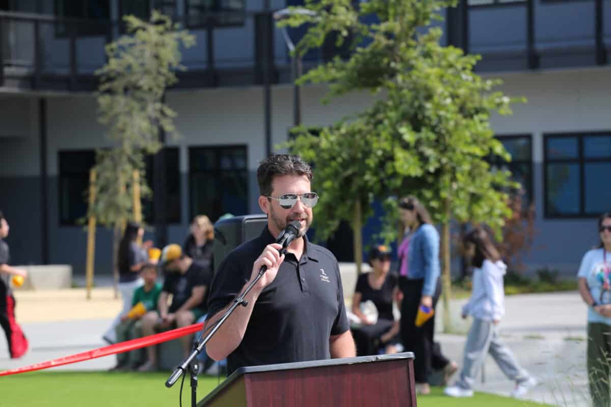 Alameda post - A man in a polo shirt and sunglasses speaks into a microphone while outdoors on the new Wood Middle School campus.