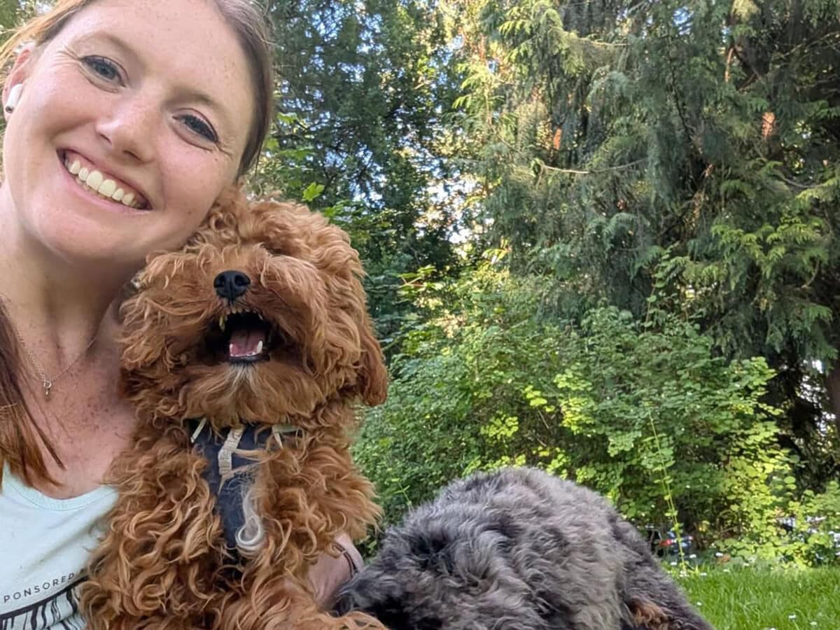 Alameda Post - Catie Norboe smiles outdoors in a park or backyard with two small fluffy dogs.