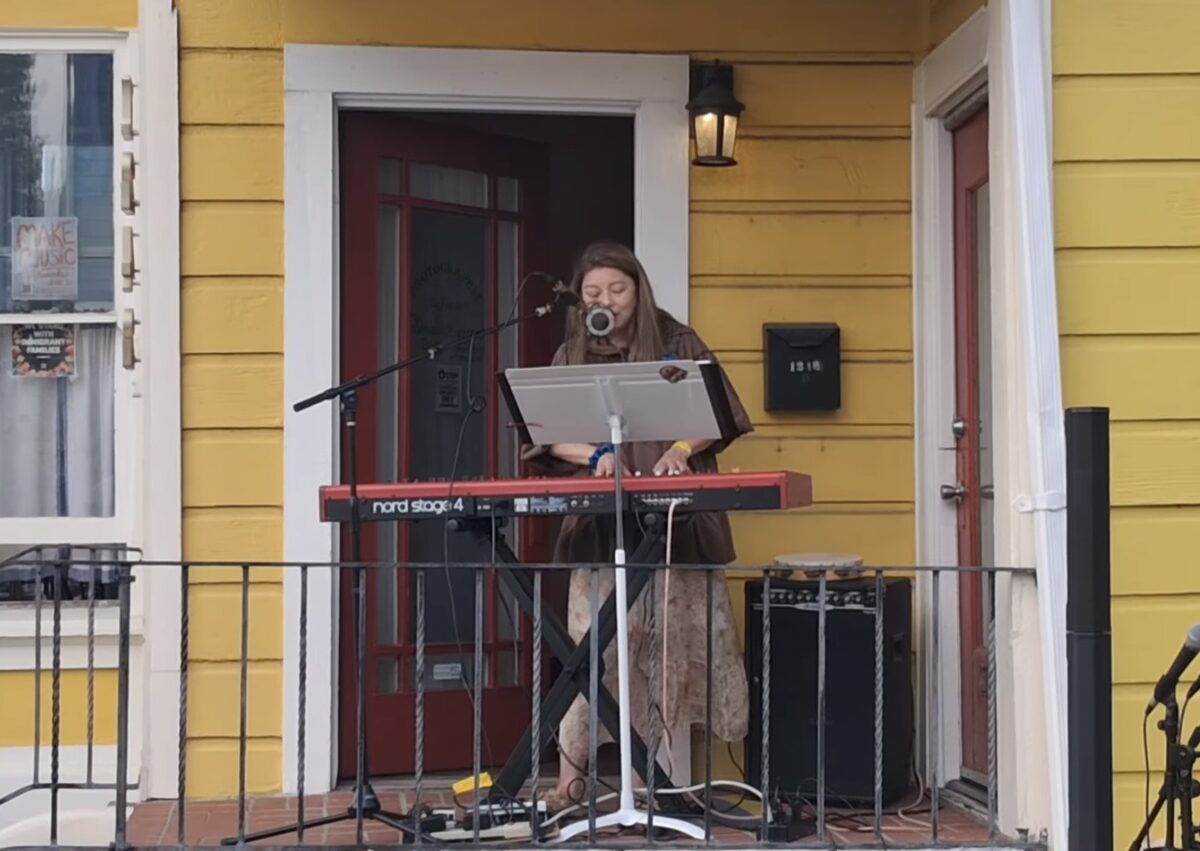 Alameda Post - A woman plays the keyboard and sings into a microphone on a porch. 