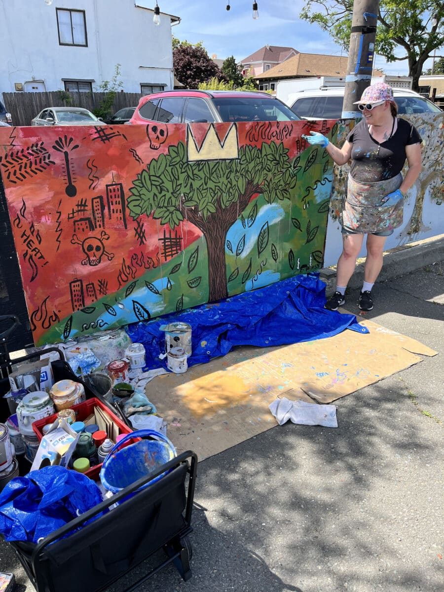 Alameda post - An artist stands next to a mural of a tree with a crown, amid a juxtaposed background of destruction.