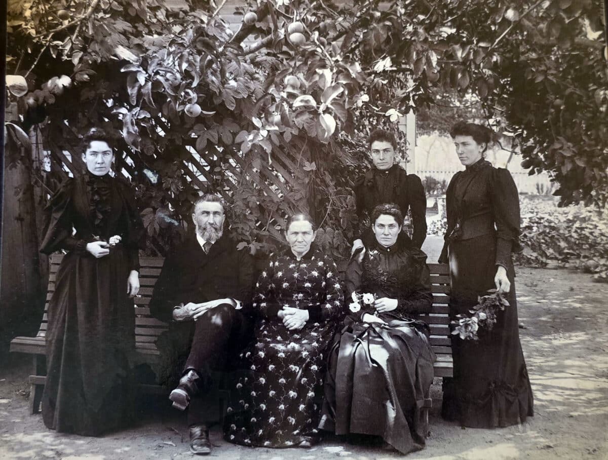 Alameda Post - A formal portrait of two parents and fourth adult daughters standing and sitting on a bench in an outdoor space. They all wear dark colors.