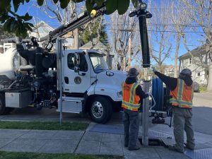 Alameda Post - two workers in bright orange vests maneuver a large pipe connected to a truck into the ground under a grate