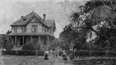 Alameda Post - A beautiful black and white photo of a family in front of a large victorian era home, hedges, hard, and trees.