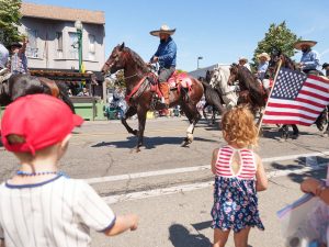 Alameda Post - A young girl wearing a red, white, and blue dress waves an American flag while watching horses in the fourth of July parade.