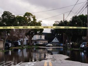 Alameda Post - A piece of caution tape across an intersection. Flooding on an Alameda street is visible in the background