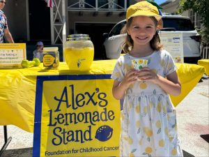 Alameda Post - A young girl wearing a dress with lemons on it stands in front of a lemonade stand. She holds a cup and smiles.
