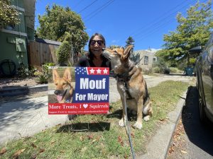 Alameda Post - Mouf the dog gives a kiss to a woman. They sit or crouch together next to a Mouf for Mayor yard sign by the road.