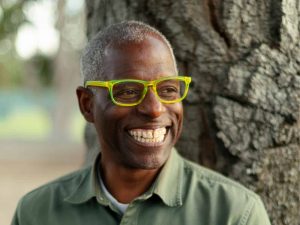 Alameda Post - Amos White smiles in front of a tree wearing bright yellow rimmed glasses