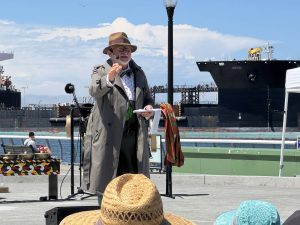 Alameda Post - A man in a hat and trench coat performs a skit in front of the Oakland Alameda Estuary
