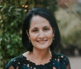 Alameda Post - Nancy Lee Bronstein, a woman with a black bob hairstyle, smiles at the camera in a beautiful headshot. She is in an elegant top that may be a dress, and in a garden.
