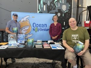 Alameda Post - Three people stand and sit around an Ocean Hoptimism booth. Two of them hold a book titled Forest of the Sea.