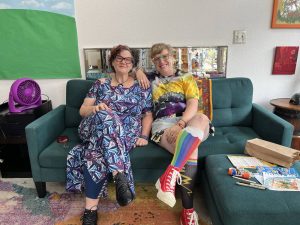 Alameda Post - Two individuals in fun and colorful clothes sit on a couch together and smile. They are two of the owners of Craftwork.